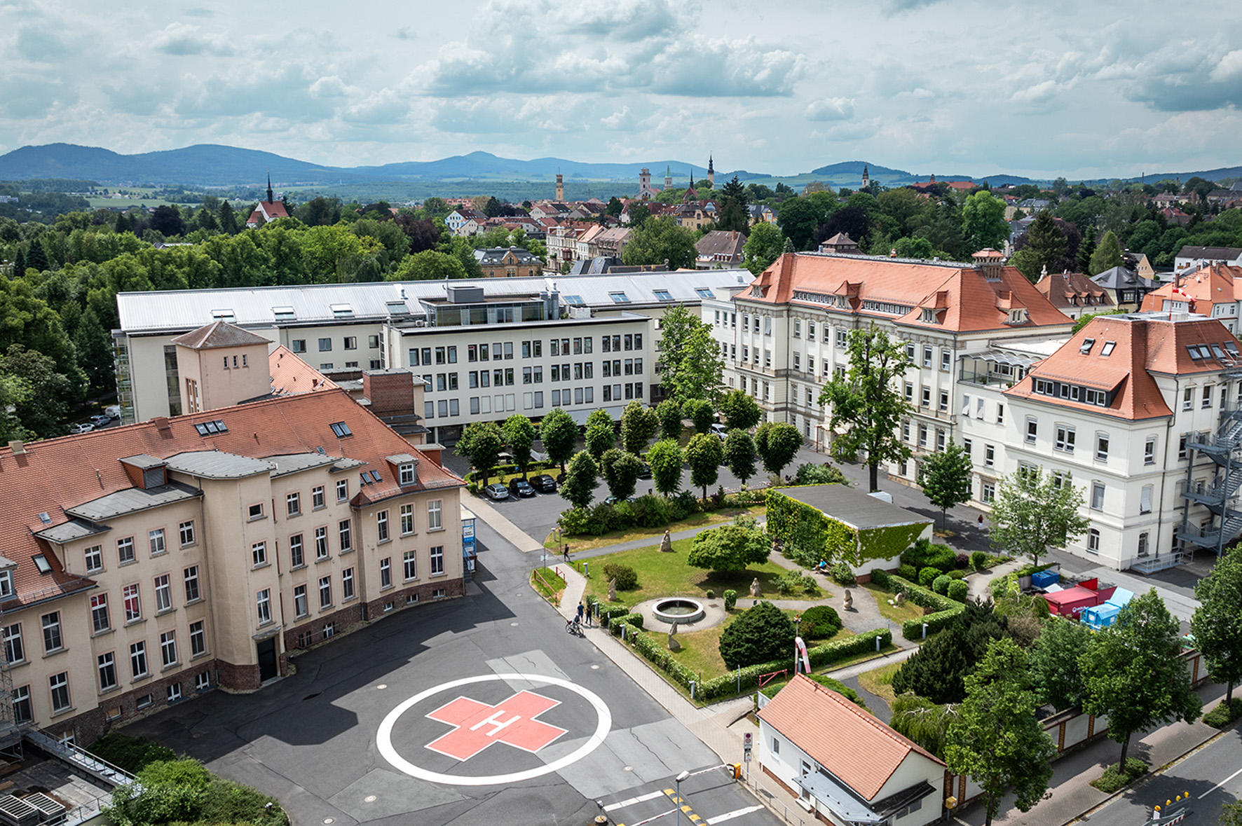 Eine Luftbildaufnahme des Krankenhauses in Zittau. Im Vordergrund ist die Haupteinfahrt mit dem Hubschrauberlandeplatz und angrenzenden Innenhof zu sehen. Hinter dem Krankenhaus sind Häuser und Kirchtürme der Stadt Zittau erkennbar. In der Ferne schweift der Blick auf das Zittauer Gebirge bei wolkigem Himmel.