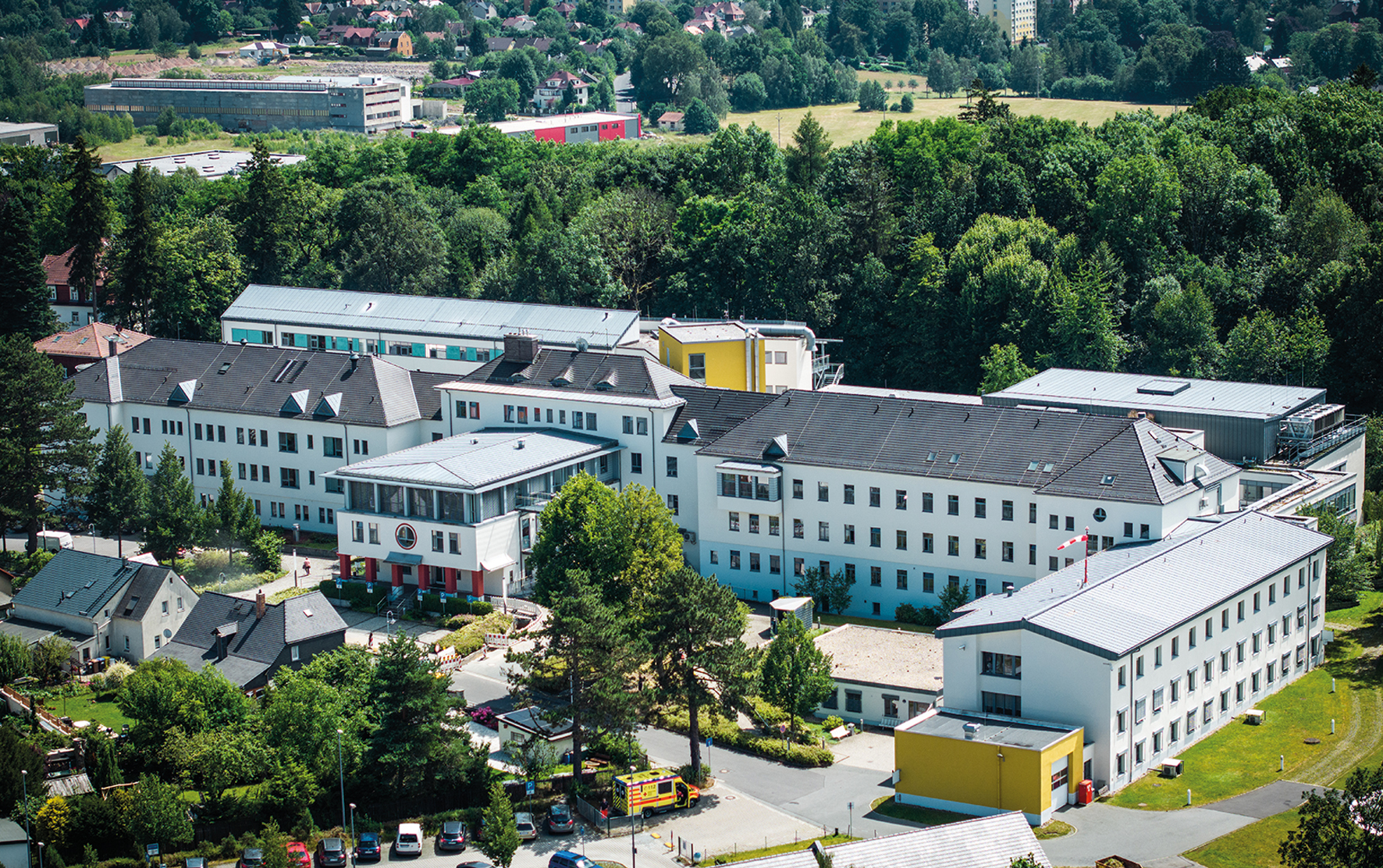 Eine Luftbildaufnahme des Krankenhauses in Ebersbach-Neugersdorf. Im Vordergrund sind ein Parkplatz und angrenzende Wohnhäuser zu sehen. Der Blick auf das Krankhaus richtet sich aus Richtung der Notaufnahme auf den Haupteingang. Auch die Bettenhäuser sind zu sehen. Hinter dem Krankenhaus grenzt ein Waldstück an.