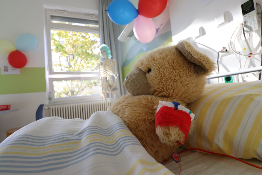 Ein Teddybär liegt in einem Krankenhausbett, der Arm ist verbunden. Im Hintergrund ist das Patientenzimmer mit Luftballons geschmückt